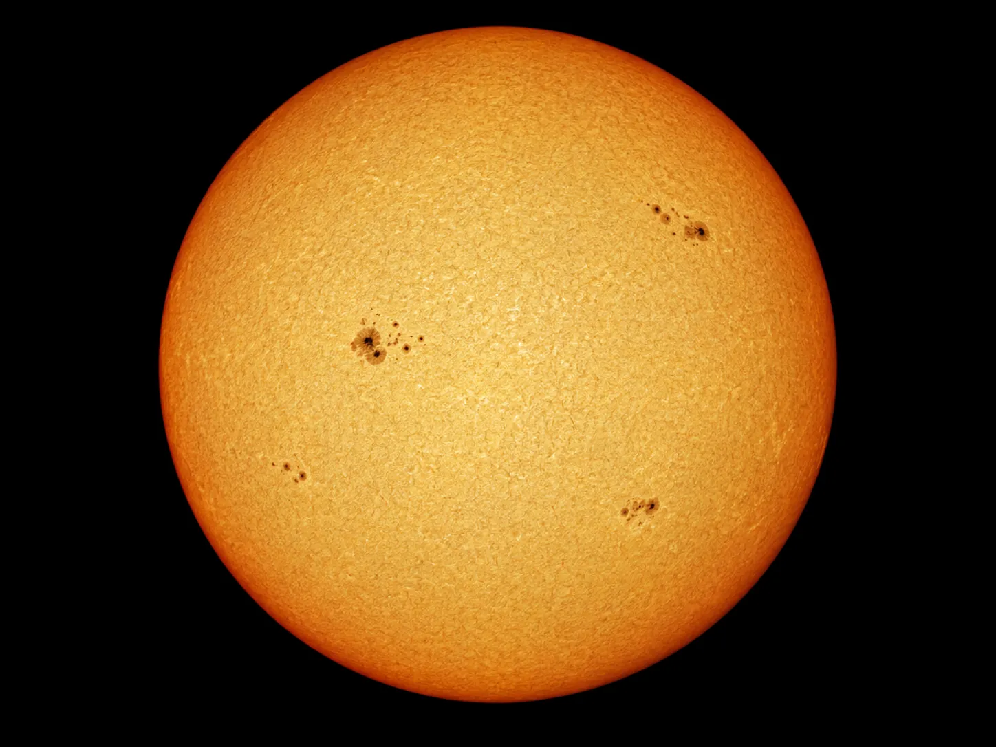 太陽観察用のフィルターを通して見た太陽全体の実写風の画像。だいだい色に光る球で、ふちほど暗く、表面に黒い点(黒点)のあつまりが4か所ある